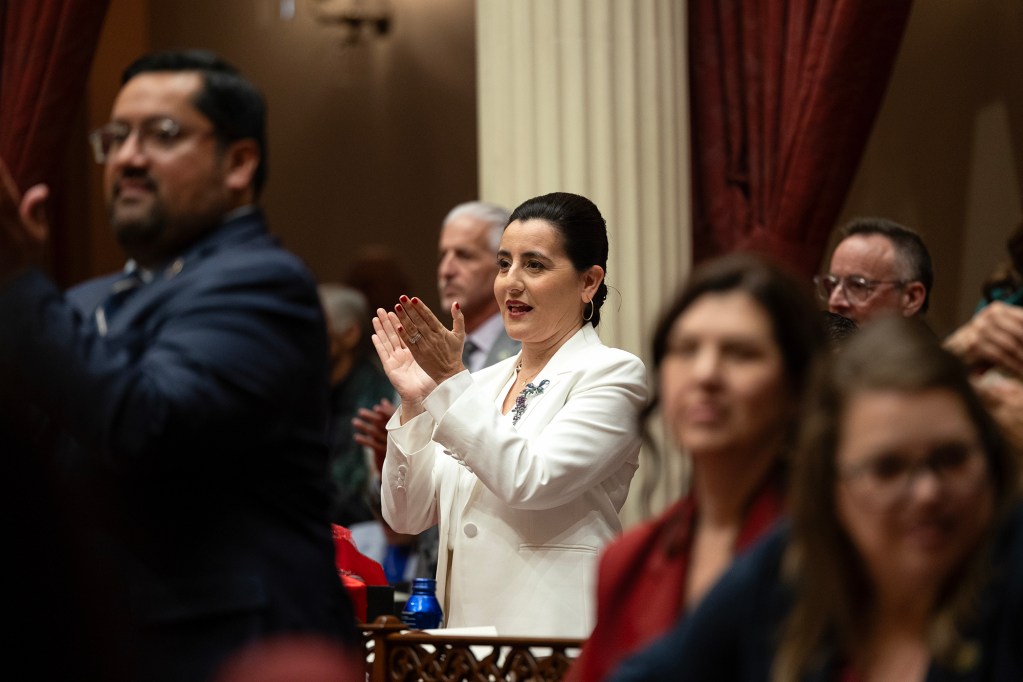 A person in a white suit applauds while standing among colleagues inside a legislative chamber, as others around them also stand and clap. The scene is framed by tall columns, red drapes, and warm interior lighting, with several people partially visible in the foreground and background.
