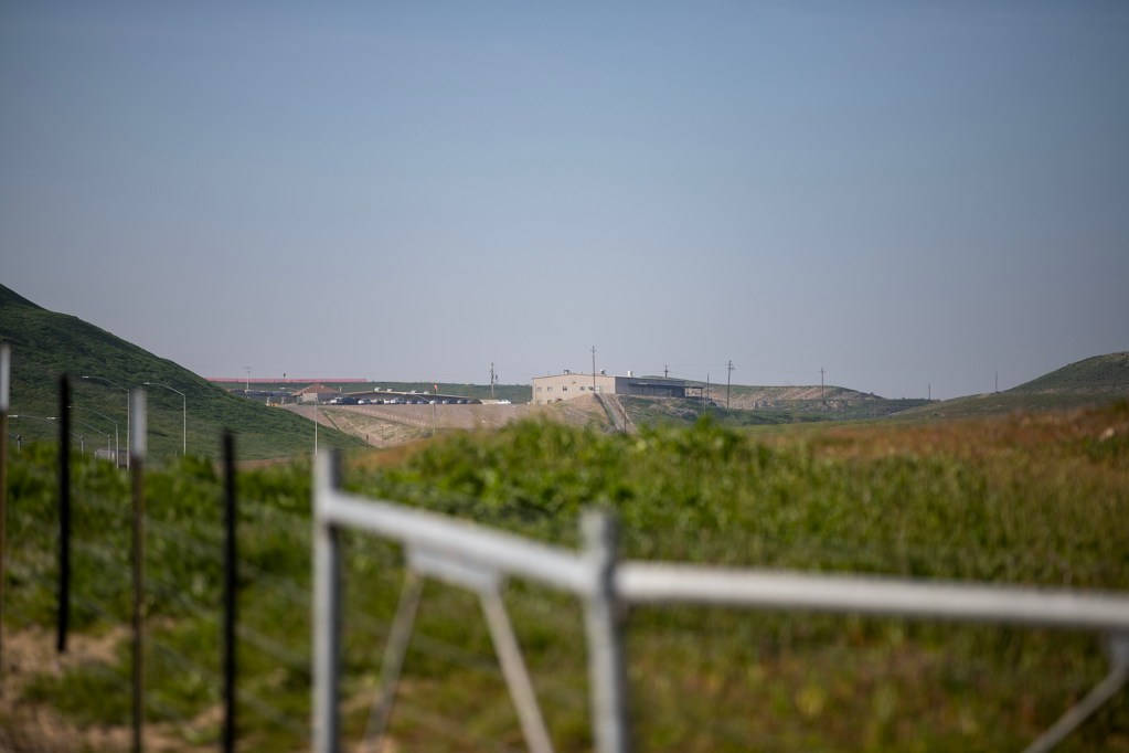 A distant industrial facility sits on a low ridge between rolling green hills, seen beyond a blurred fence and grassy foreground under a pale blue sky.