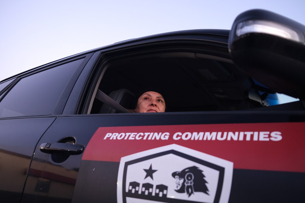 A person, sitting on the passenger seat of a car, looks out the window. A large sticker with the words "protecting communities" is placed on the outside of the passenger's door.
