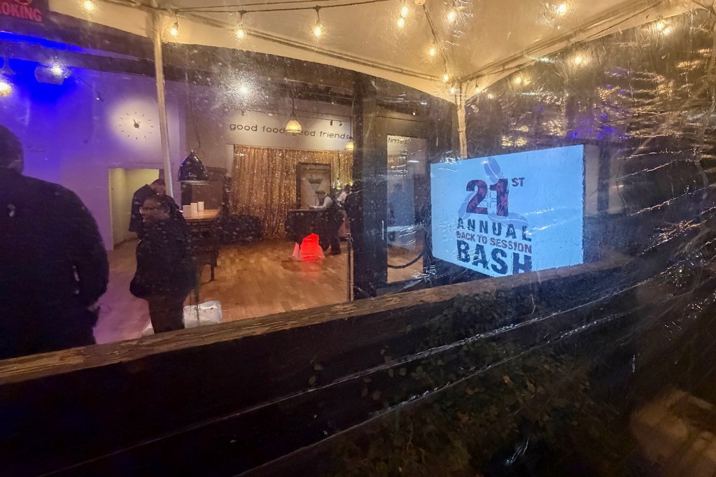 View through a clear plastic tent wall into a warmly lit indoor space with string lights overhead, people mingling inside, and a sign reading “21st Annual Back to Session Bash” visible on a screen.