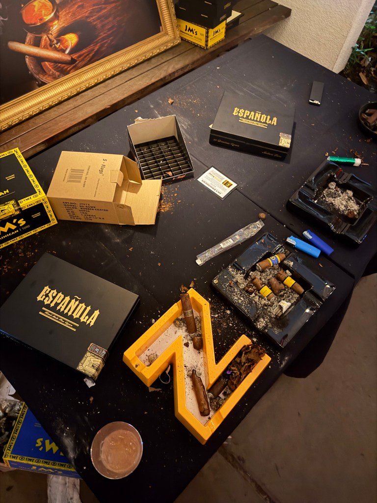 Overhead view of a black tabletop scattered with cigar boxes, loose cigars, ashtrays filled with ash and cigar ends, lighters, and cigar debris, including a yellow letter-shaped tray holding partially smoked cigars.
