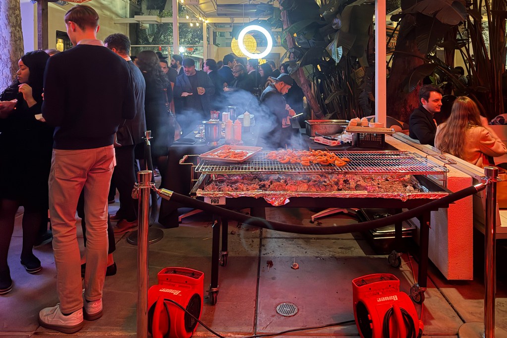 An outdoor nighttime gathering with a large grill cooking skewers and meat in the foreground as smoke rises, while a crowd mingles around tables and food stations under string lights and patio heaters.
