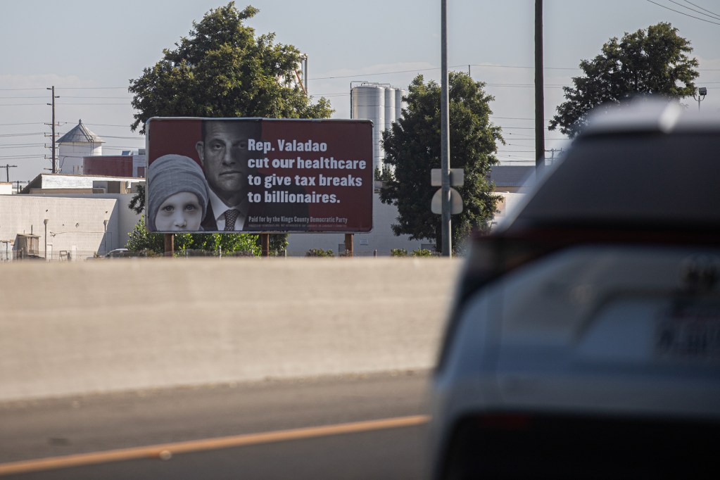 A red political sign is seen near a highways with cars passing through. That sign, that includes an image of a lawmaker next to an image of a child, reads " Rep. Valadao cut our healthcare to give tax breaks to billionaires."