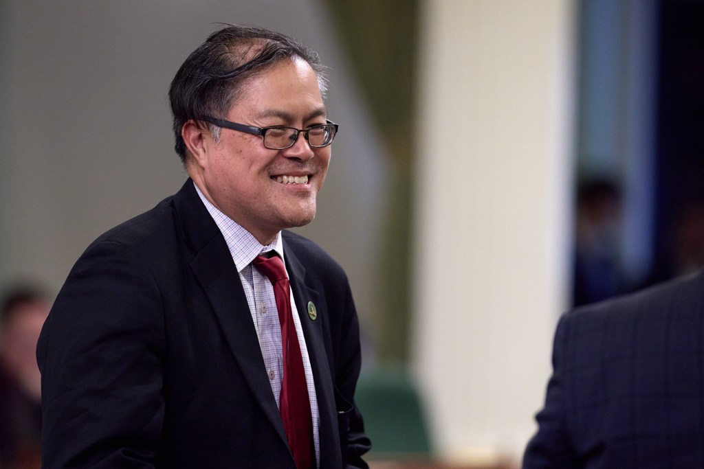 A close-up view of a lawmaker wearing a black suit and red tie as he smiles and looks towards another person off-frame.