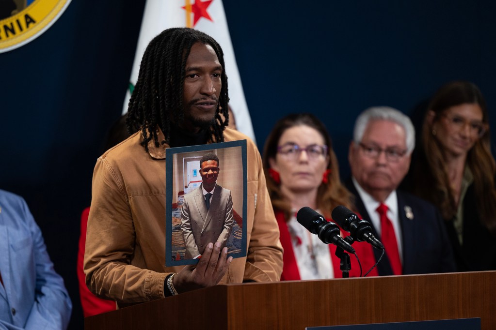 A person stands at a podium holding a framed photo of another person while speaking into microphones, with several people seated behind them and flags in the background.