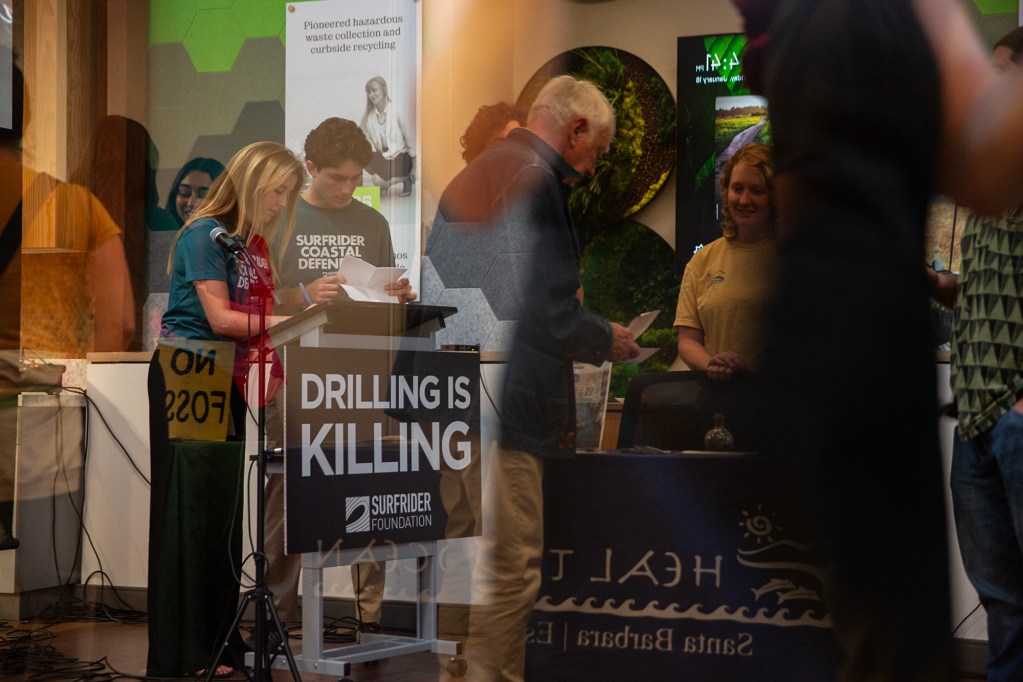 The reflection of people as they stand and mingle during a town hall. Two of those people stand behind a podium with a sign that reads, "drilling is killing" as they look at various sheets of paper.