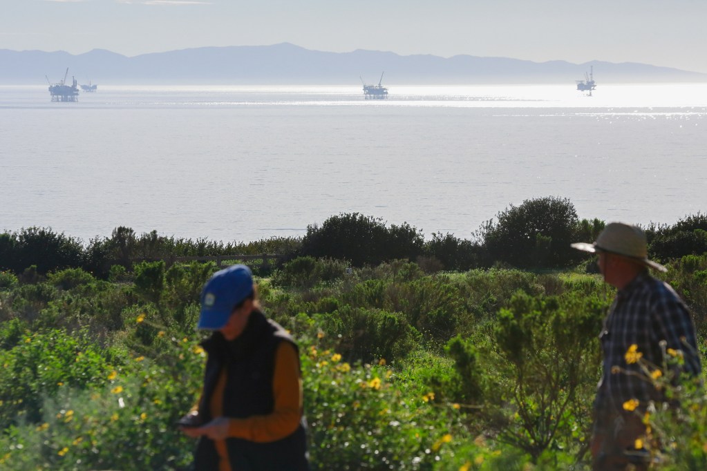 Oil rigs positioned in the middle of the ocean can be seen in the far distance as two people, in the foreground of the photo, walk through a path surrounded by greenery.