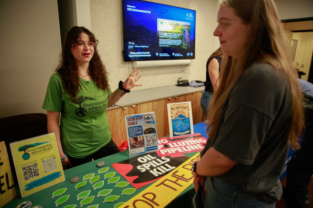A person wearing a gray shirt listens to another person talk as they stand in front of a table with signage and stickers related to oil spills.