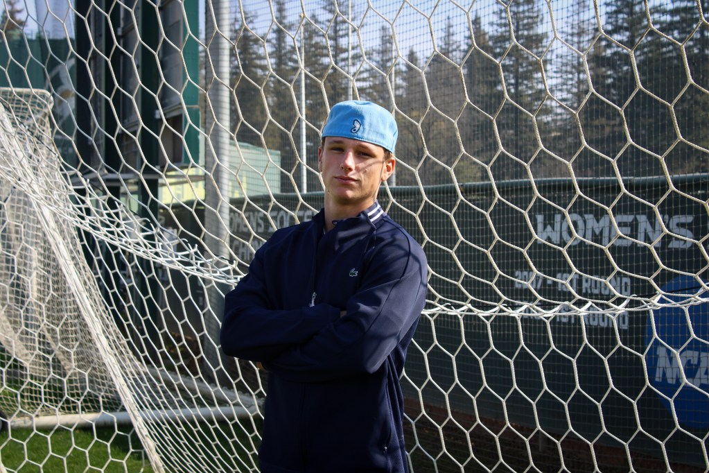 A person stands with arms crossed in front of a soccer goal net on an outdoor field. They wear a light blue cap and a dark athletic jacket, looking toward the camera with a composed expression. The netting fills most of the background, with trees and fencing visible beyond.