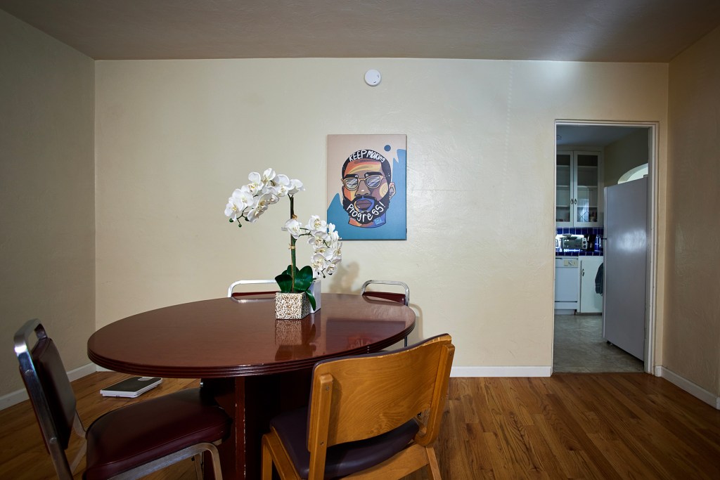 A small dining area features a round wooden table with mixed chairs and a potted white orchid centerpiece. A notebook rests on one chair, and a framed illustration hangs on the plain wall above. At right, an open doorway reveals part of a kitchen with white cabinets and blue tile counters, while wood flooring runs throughout the room.
