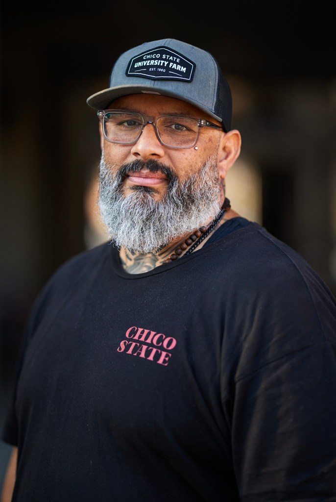 A person with a gray beard and mustache looks toward the camera with a slight smile. They wear glasses, a dark cap that reads “Chico State University Farm,” and a black T-shirt with “Chico State” printed on the chest. A beaded necklace is visible at their collar, and faint tattoos appear on their neck. The background is softly blurred and dark, drawing attention to their face.
