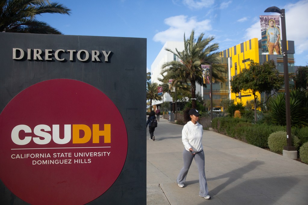 A person walks through a walkway near a directory sign with the California State University Dominguez Hills logo on it. Palm tress and a yellow building can be seen in the distance.