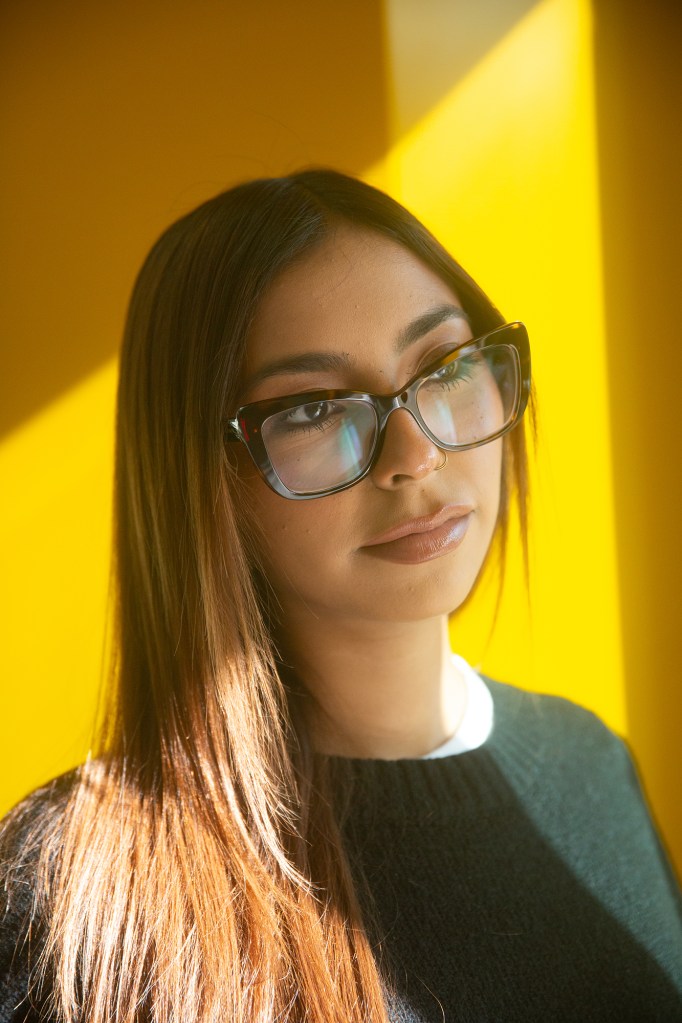 The sunlight softly illuminates the right side of a person as they look off into the distance and stand near a window with a yellow wall behind them. The person has long brunette hair and wears reading glasses.