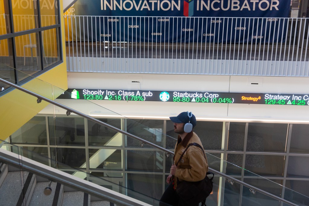 A person wearing headphones walks up a flight of stairs as a LED ticker shows different stocks.