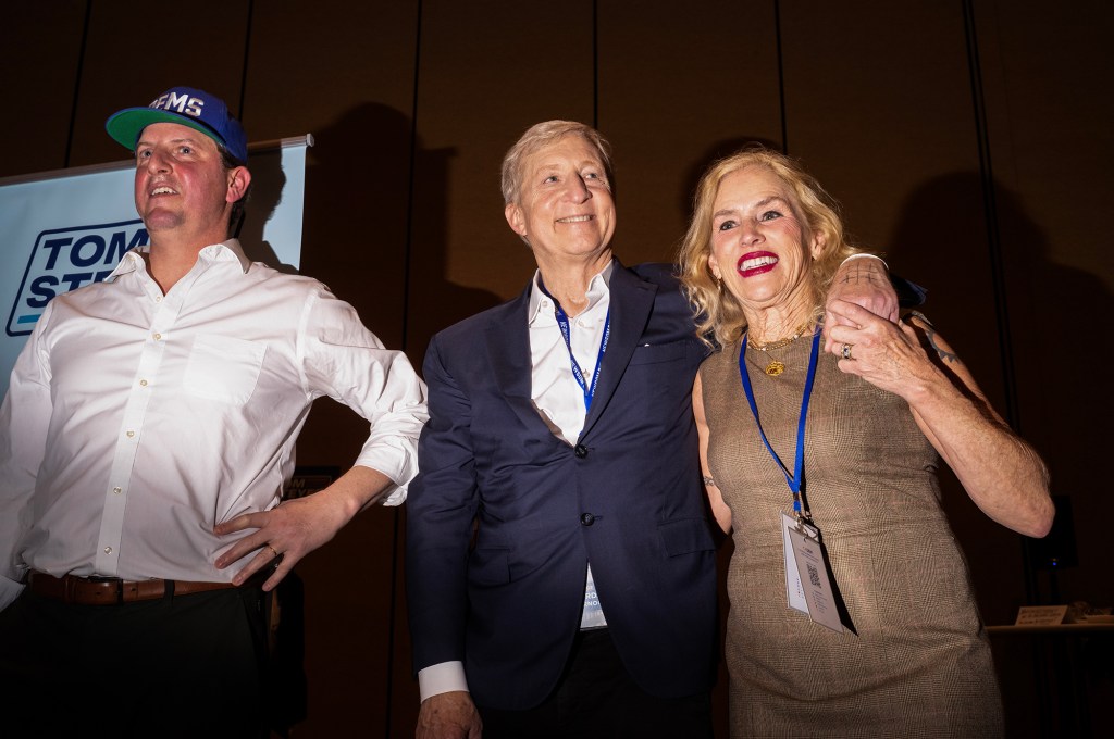 Three people stand closely together indoors at an event, smiling and posing with their arms around each other. The person in the center wears a dark suit and lanyard, the person on the right wears a sleeveless dress with a lanyard badge, and the person on the left wears a white shirt and a blue baseball cap. A sign partially visible behind them suggests a political or conference setting.
