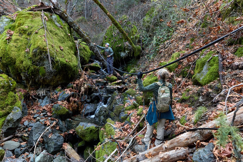 Two people, both dressed in hiking gear and carrying backpacks, climb along rocks in a small river, holding a black plastic hose that guides them along the creek.