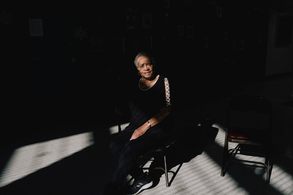 A person sits on a chair in a dim room, illuminated by a beam of light from a window, with another empty chair nearby.