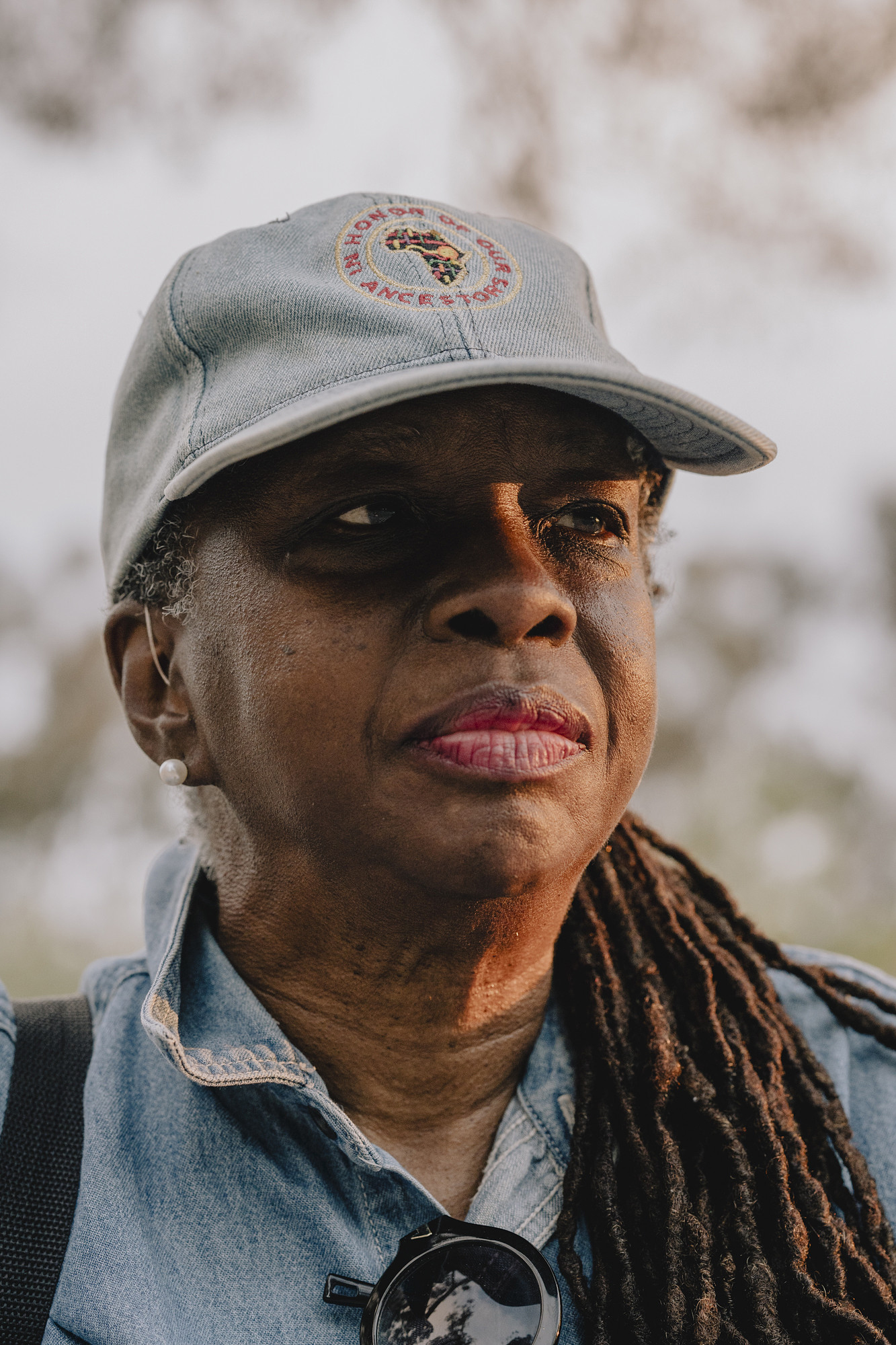 A close-up of a person wearing a baseball cap, earrings, and a denim shirt, looking off to the side with trees blurred in the background.