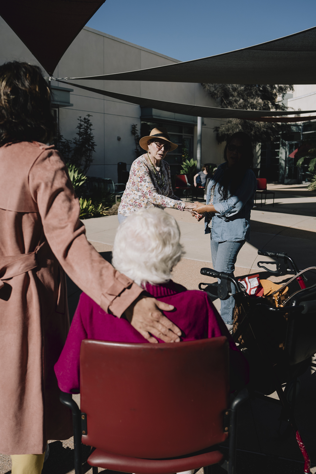 In an outdoor courtyard, a seated person with white hair faces two people shaking hands while another person stands beside the chair with a hand resting on their shoulder, and a walker is visible nearby.