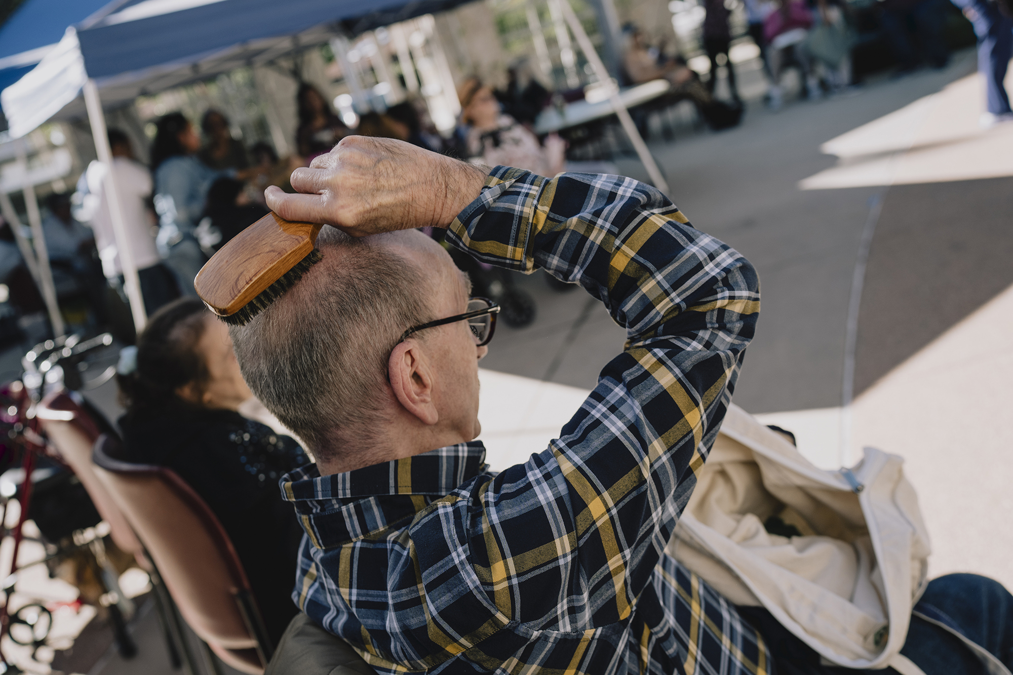 An older person in glasses and a plaid shirt sits outdoors under a canopy, brushing the top of their head while others gather in the background.