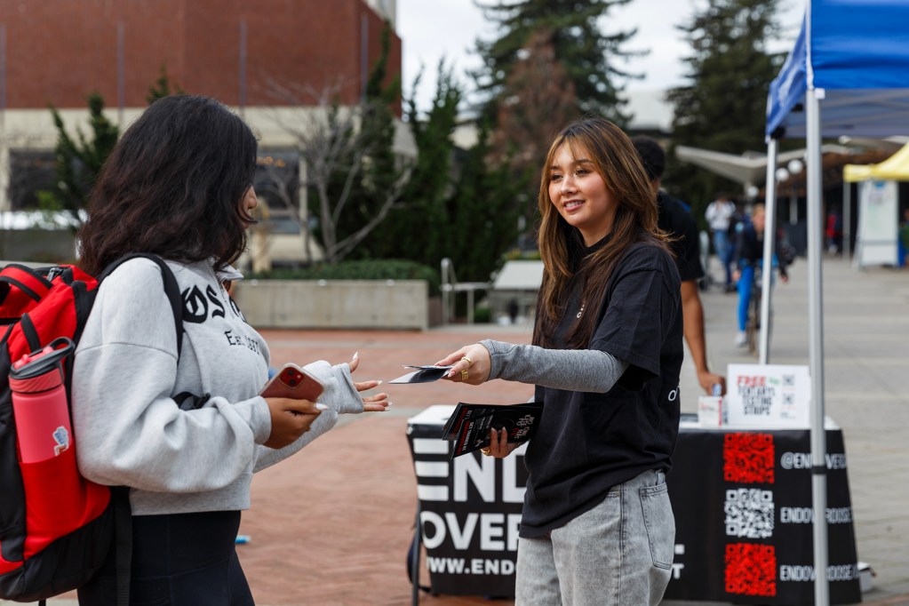A person hands a flyer to another person at an “End Overdose” outreach table set up in a campus plaza.