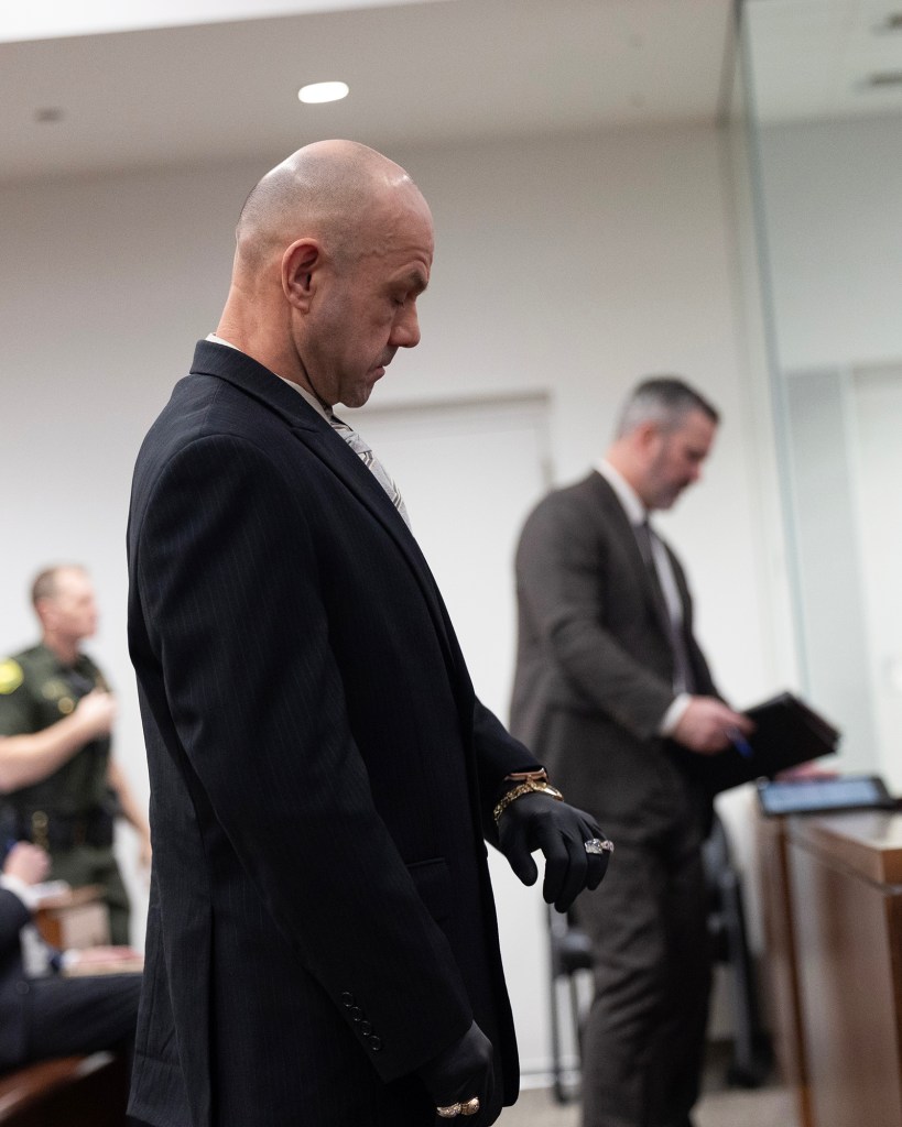 A person in a suit and black gloves stands in a courtroom with head slightly lowered, while another suited person stands at a lectern in the background. A uniformed officer and other attendees sit further back beneath bright ceiling lights.