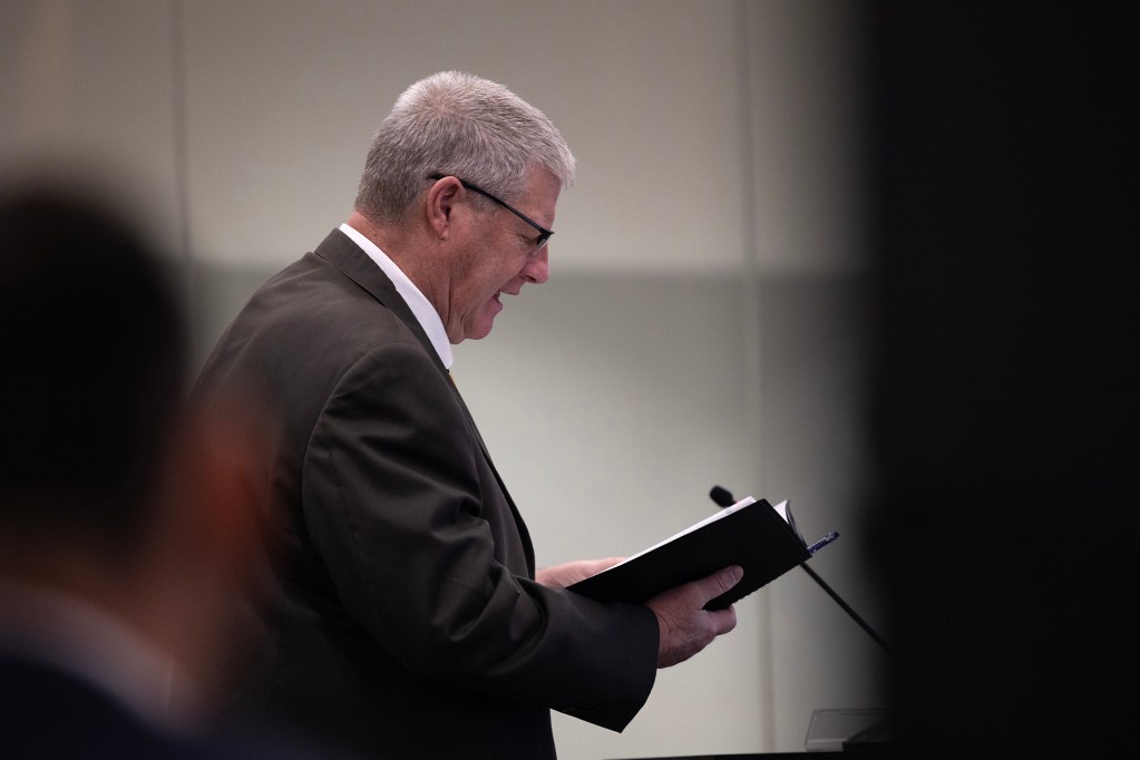 A person in a suit stands at a microphone reading from a notebook during a court proceeding, viewed from the side. The background is softly lit and out of focus.