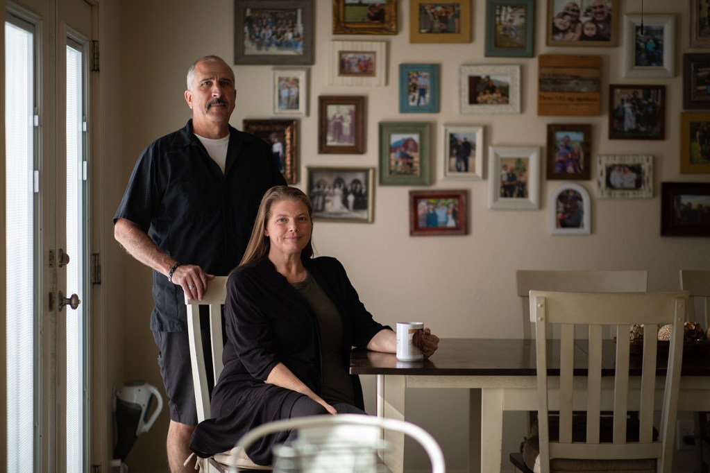 A person stands behind another person sitting to pose for a photo in a dining room area near a kitchen table. In the background is a plain white wall with an assortment of family photos hanging on it.