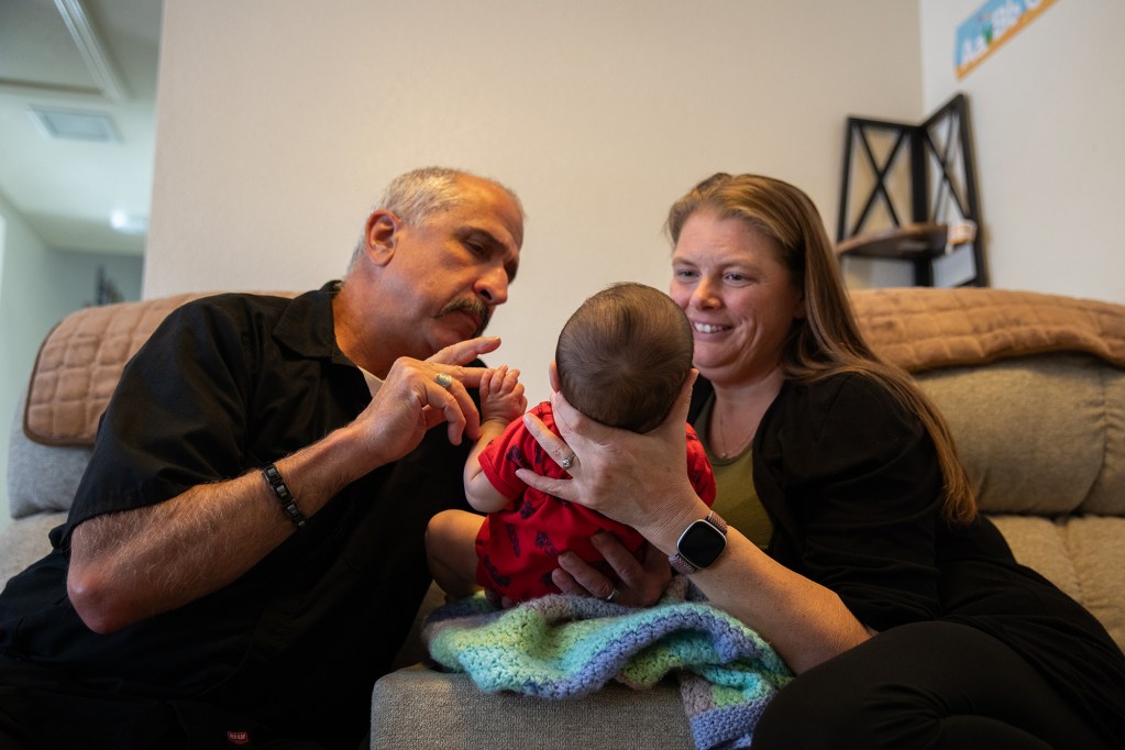 Two people sitting on a couch, holding a baby, are supporting the baby's neck so the baby can look and play with it. In the background is the set of a family’s living room.