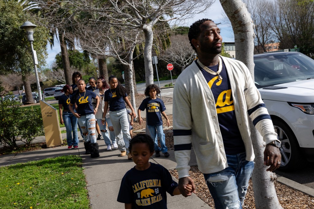 A person wearing a white sweater with blue stripes holds a young child's hand as they walk along a sidewalk near a park. Behind them is a group of young children wearing matching blue shirts with the UCLA logo.