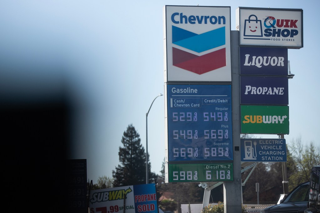 A Chevron gas station sign displays fuel prices for regular, plus, supreme, and diesel. Additional signs advertise a convenience store, liquor, propane, Subway, and an electric vehicle charging station.