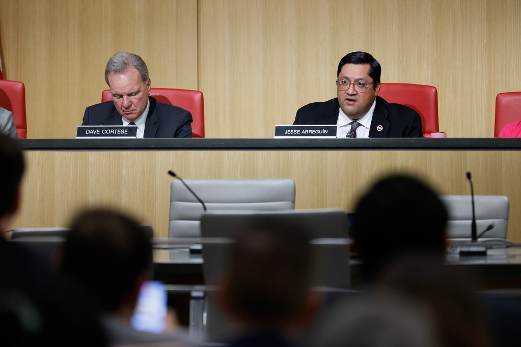 Two lawmakers sit behind a dais during a public hearing, with nameplates reading “Dave Cortese” and “Jesse Arreguín.” One looks down at documents while the other speaks, as attendees sit in the foreground.
