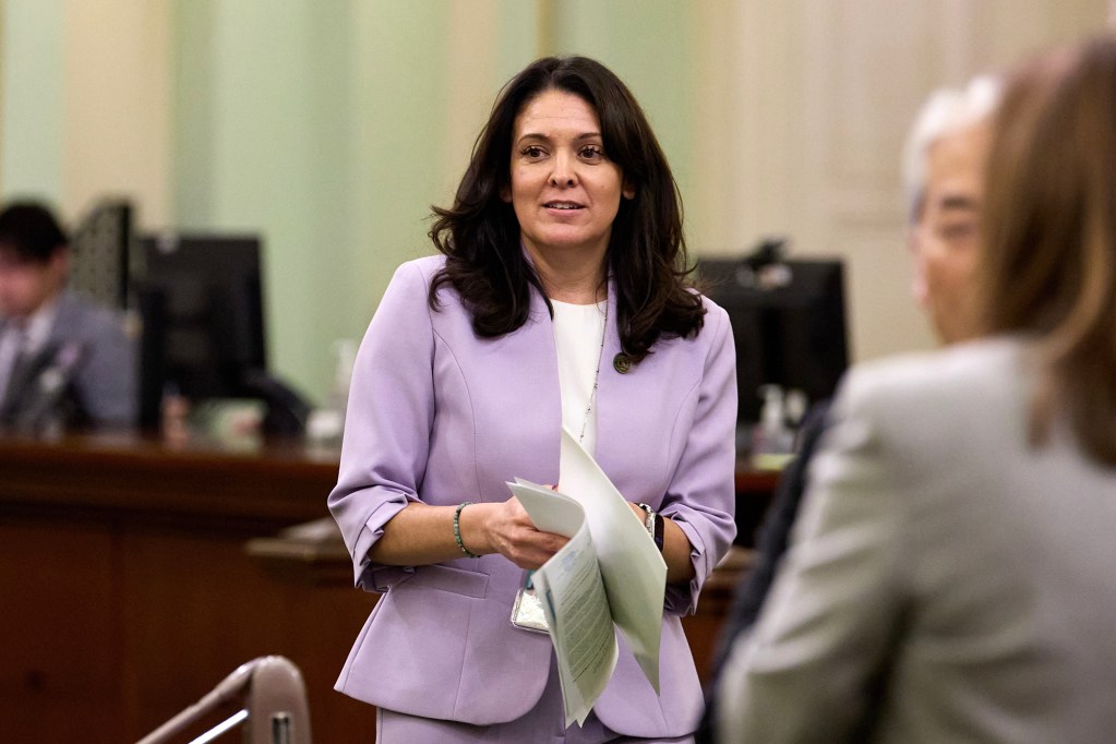 A lawmaker stands on the Assembly floor holding a stack of papers, speaking with colleagues during a legislative session. The chamber’s desks and computer monitors fill the background as other lawmakers sit and confer nearby.