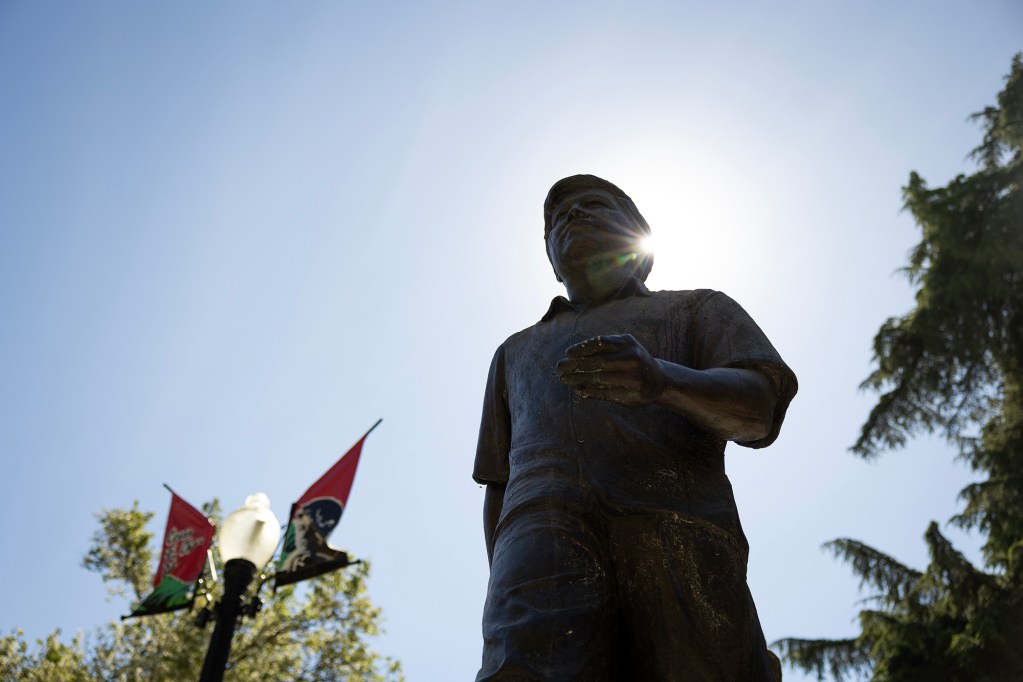 A low-angle view of a bronze statue of a person standing with one hand slightly extended, silhouetted against a bright sky. Sunlight flares from behind the figure’s head, while a streetlamp with red and green banners and leafy trees frame the scene below.