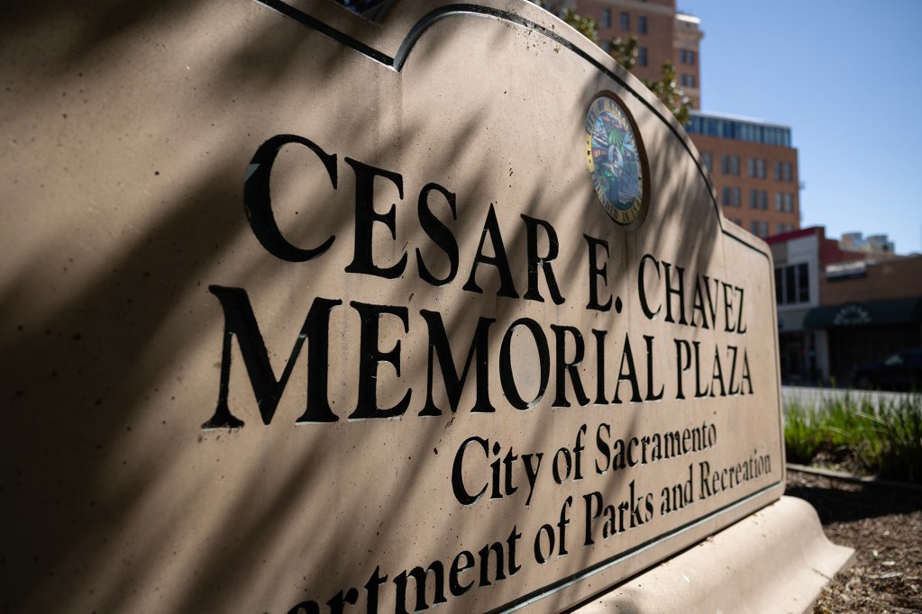 A stone sign reading “Cesar E. Chavez Memorial Plaza” stands in a landscaped urban area, with smaller text noting the City of Sacramento Department of Parks and Recreation. Sunlight casts soft shadows across the engraved lettering, while nearby buildings and greenery appear in the background.