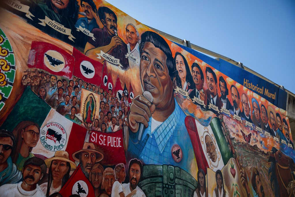 A large, colorful mural covers a building wall, featuring a central figure holding a microphone surrounded by portraits of activists and historical figures. Crowds, flags and text such as “Si Se Puede” fill the scene, with the composition blending portraits, protest imagery and cultural symbols under a clear blue sky.