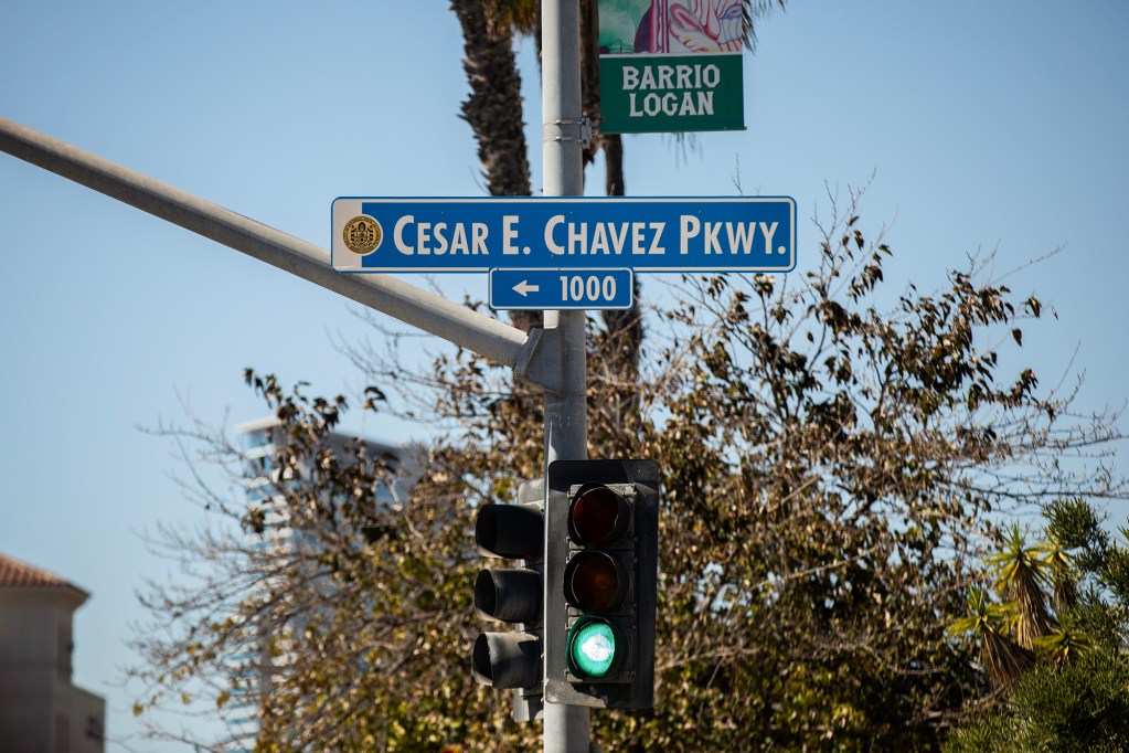 A blue street sign reading “Cesar E. Chavez Pkwy” is mounted on a traffic light pole, with a smaller sign below indicating the 1000 block. A green traffic light glows beneath it, while a “Barrio Logan” banner, palm trees and leafy branches frame the scene against a clear sky.