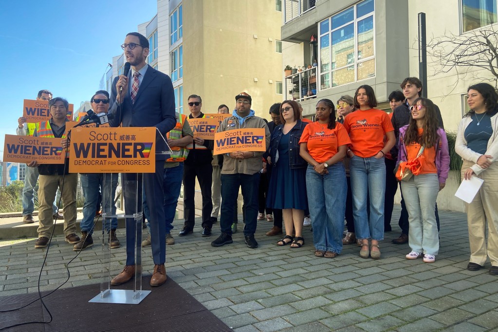 A candidate speaks at a podium during a campaign event while supporters stand behind them holding &ldquo;Scott Wiener for Congress&rdquo; signs. The group gathers outside a residential building under a clear sky.