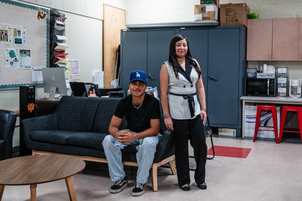 Two high-school-age students pose for a portrait. One student sits on a couch while the other stands nearby.