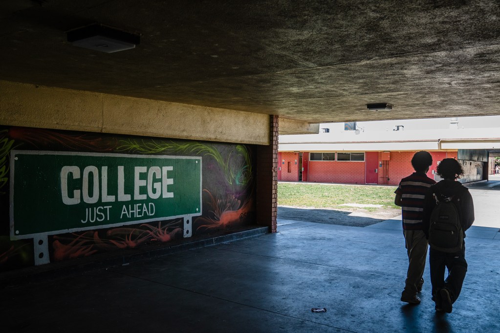 The shadow of two students walking pass a mural of a street sign named "College just ahead."