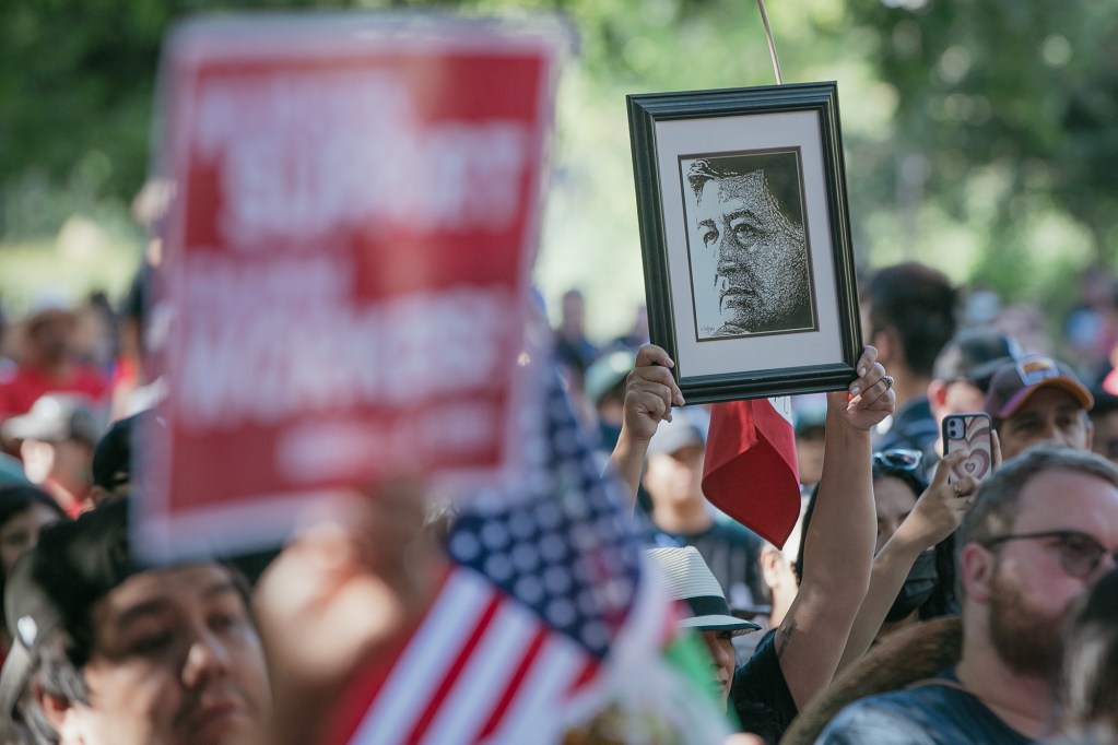 A person in a crowded outdoor gathering holds up a framed black-and-white portrait above their head. Around them, a dense group of people stands close together, some holding signs and small American flags, while others record on their phones. The background is softly blurred with trees and daylight, emphasizing the raised portrait as a focal point amid the crowd.