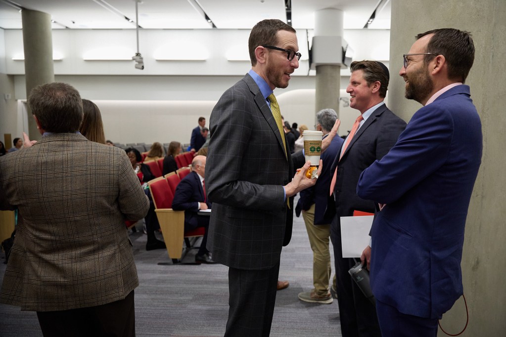 A small group of people face each other as they talk on the side of a room, where other people gather for a hearing.