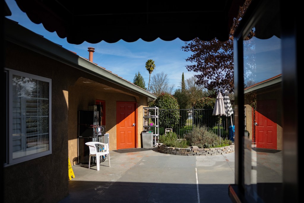 A small courtyard between buildings features red doors, a white plastic chair, and a raised garden bed with plants, enclosed by a fence under a sunny sky.