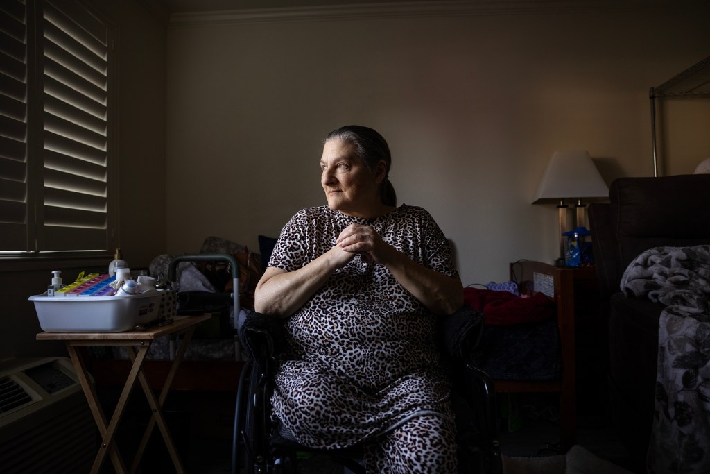 An older adult sits in a wheelchair by a window, hands clasped, looking toward light coming through blinds; a table with medications and a walker are nearby