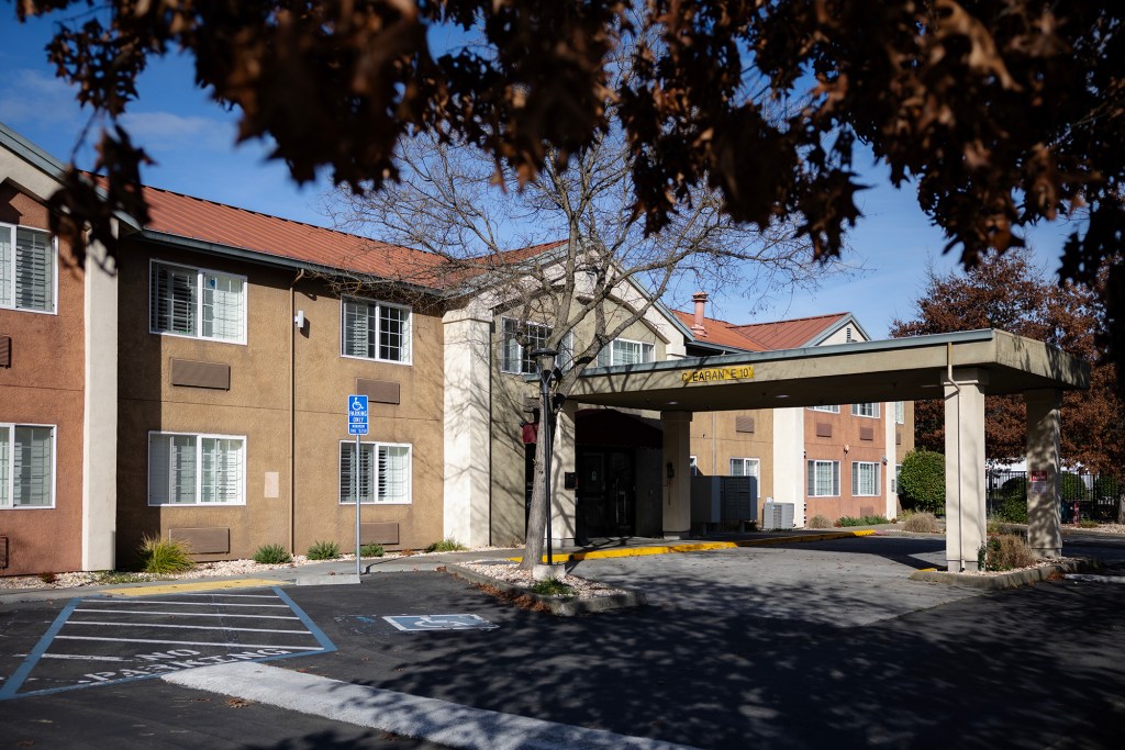 A two-story building with a covered entrance driveway sits beside a parking lot with marked accessible spaces, surrounded by trees and landscaping.