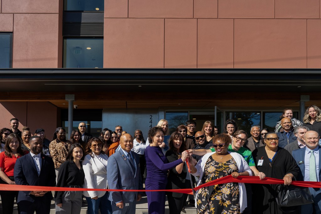 A large group of people stand outside a building entrance during a ribbon-cutting ceremony, with several individuals at the center holding scissors and cutting a red ribbon while others gather around smiling.