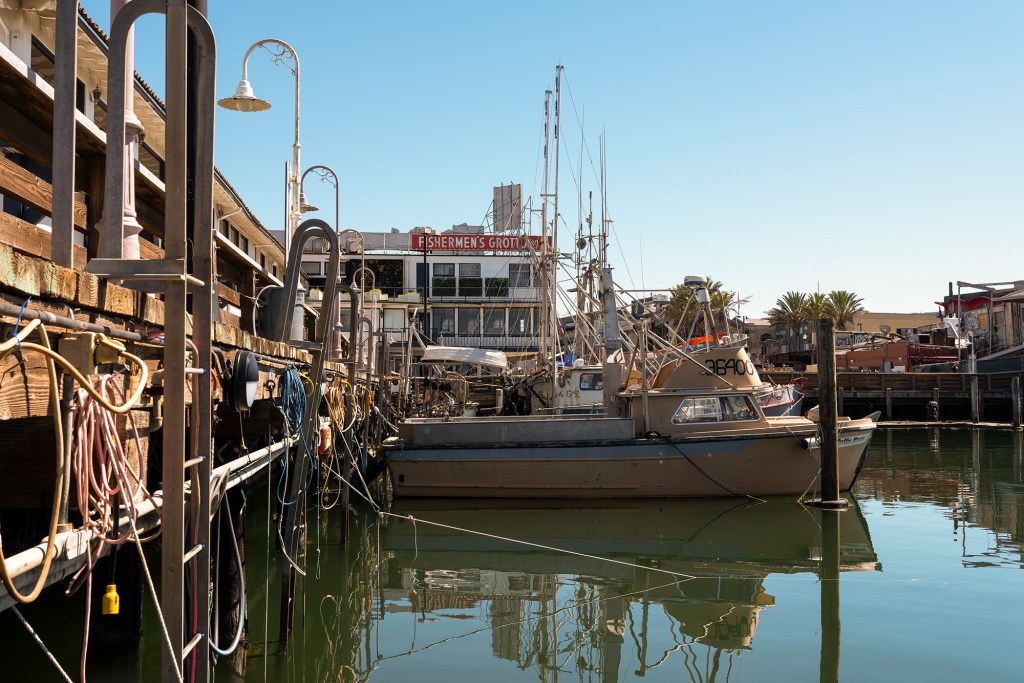 A fishing boat is moored alongside a wooden dock lined with coiled ropes, hoses, and metal fixtures. The calm water reflects the boat and dock. In the background, low waterfront buildings and masts rise against a clear sky, with a visible sign reading “Fisherman’s Grotto.”