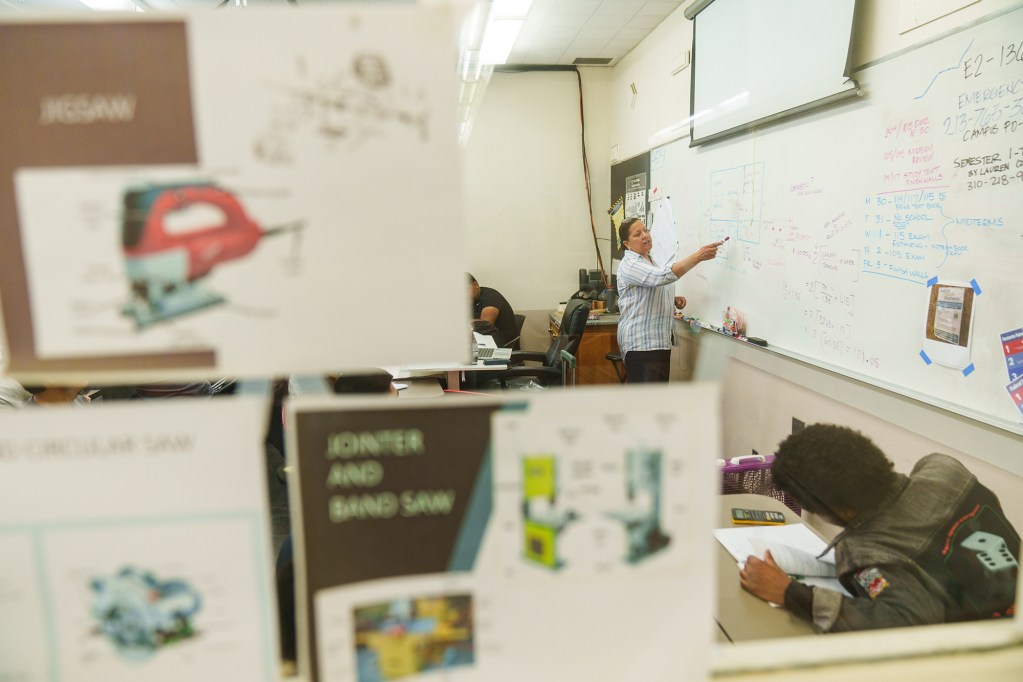 A view of a teacher teaching a lesson on a whiteboard, pointing at it with a marker while students watch, seen through a window with some paperwork taped to it.