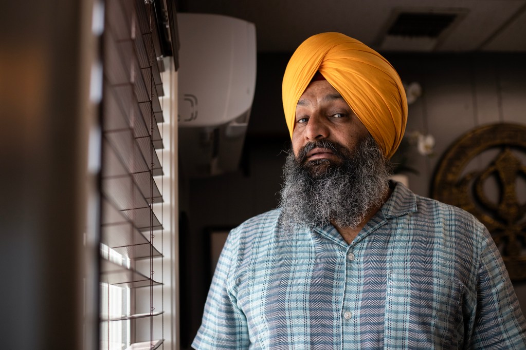 A person with a long gray beard and a bright orange turban stands indoors beside a window with horizontal blinds. Soft light from the window falls across their face, creating gentle shadows. They wear a short-sleeve patterned button-up shirt and look toward the camera with a calm, steady expression. In the background, a circular wall decoration and parts of the room’s interior are visible.