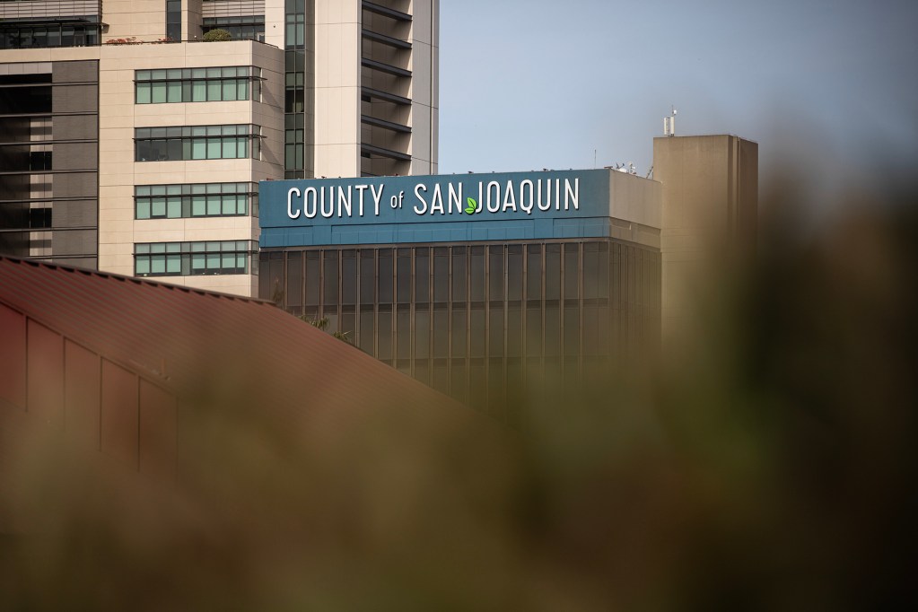 A mid-rise government building displays the words “COUNTY of SAN JOAQUIN” along its upper facade. The structure features vertical window panels and a blue band near the roofline, with taller office buildings rising behind it. Out-of-focus foliage in the foreground partially frames the lower portion of the image, adding depth.